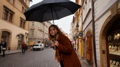 Žena s deštníkem na staré pražské ulici během deště, historická architektura v pozadí.